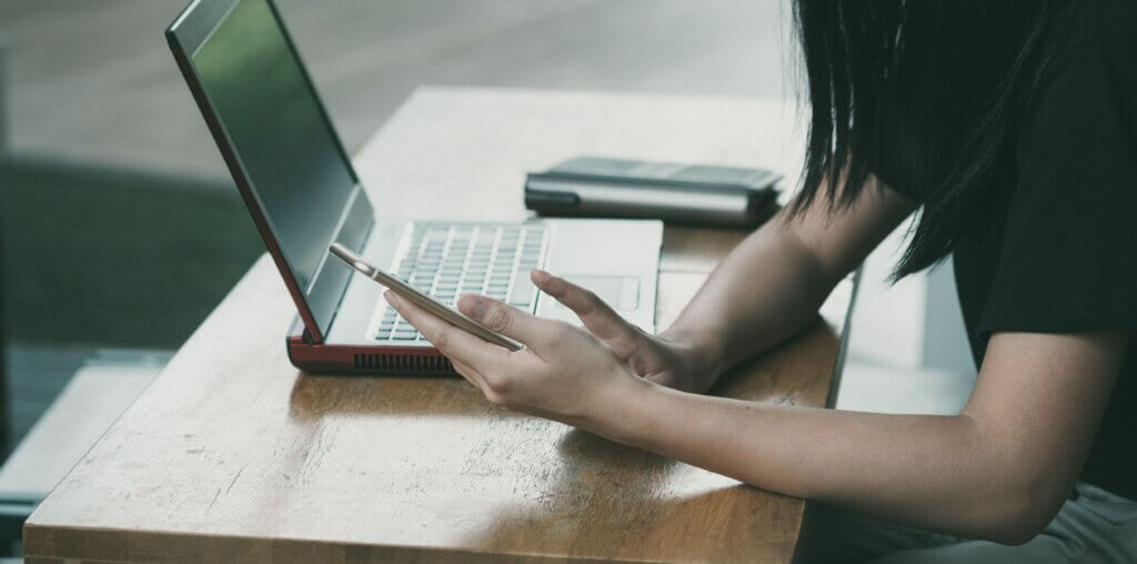 Person checking bills and account information on a smartphone while using a laptop at a desk.