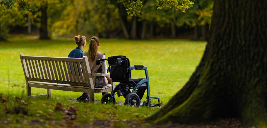Two people sit on a wooden park bench beside an empty wheelchair in a green park, illustrating accessible public seating and inclusive outdoor spaces.