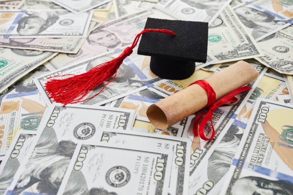 Graduation cap and diploma resting on U.S. dollar bills, representing student loans and college costs.