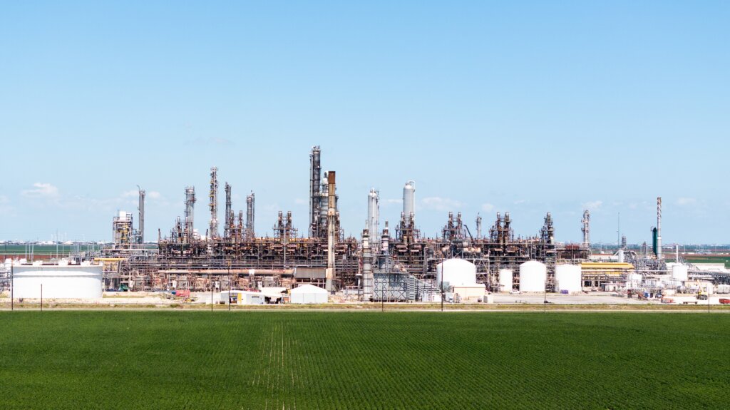 A large oil refinery with storage tanks and processing towers near Corpus Christi, Texas, stands beside open fields, representing the region's industrial and energy sector.