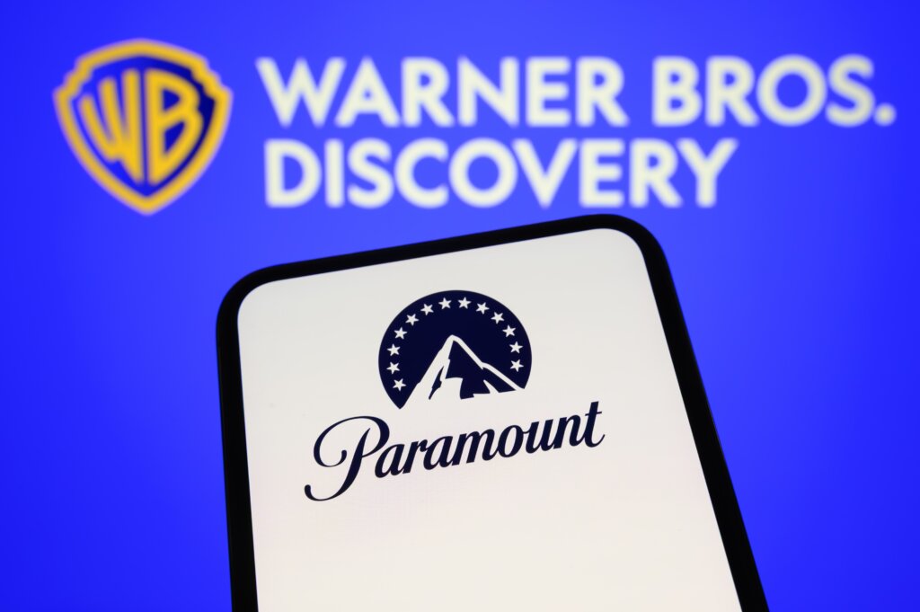The Paramount logo appears on a smartphone screen with the Warner Bros. Discovery logo displayed in the background, representing the proposed media merger.