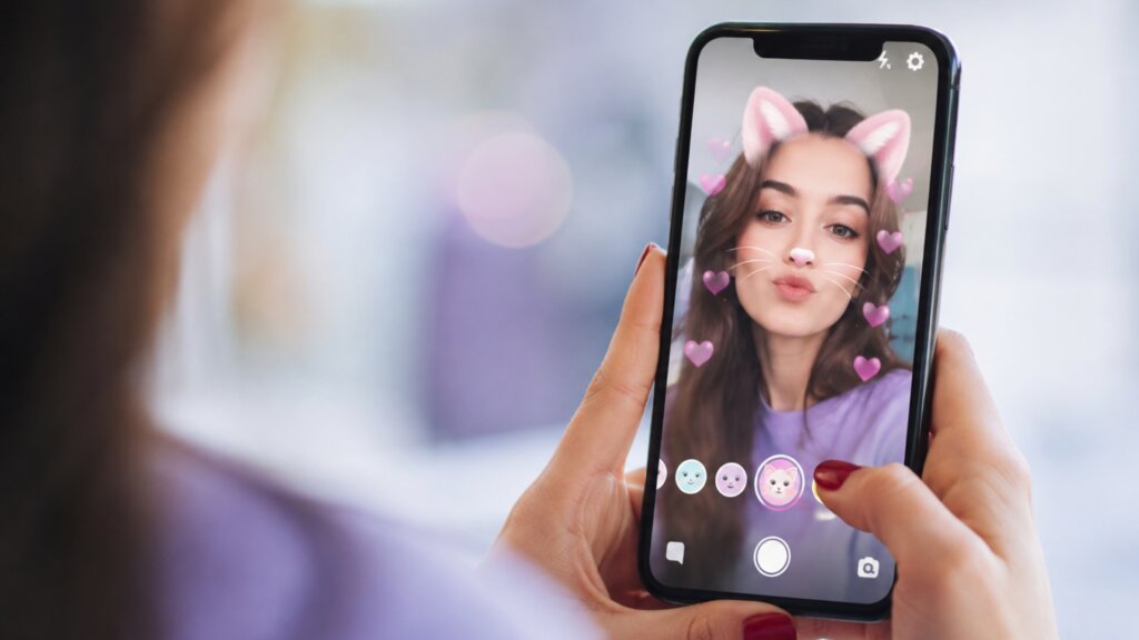 Woman using a face filter on her smartphone, with digital cat ears, whiskers, and hearts appearing on her selfie.