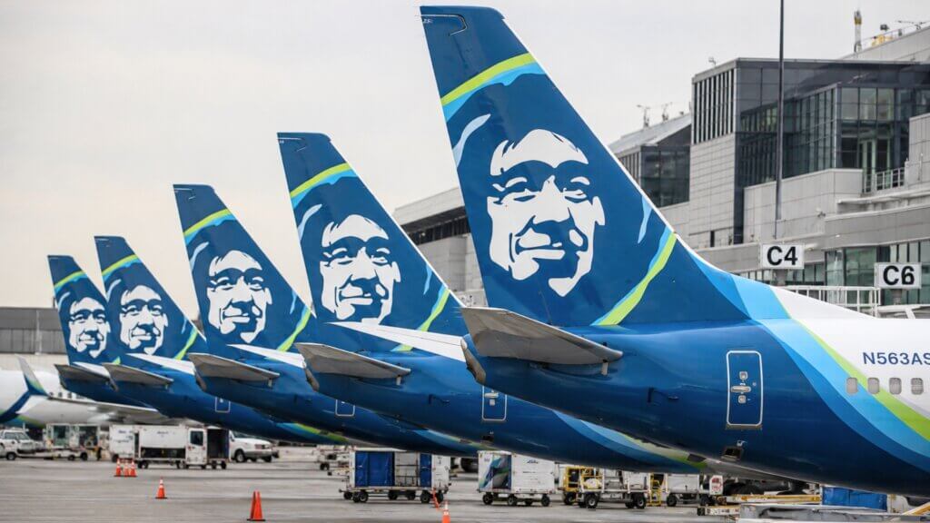 Several Alaska Airlines aircraft parked side by side at an airport gate with their blue tail fins lined up in view.