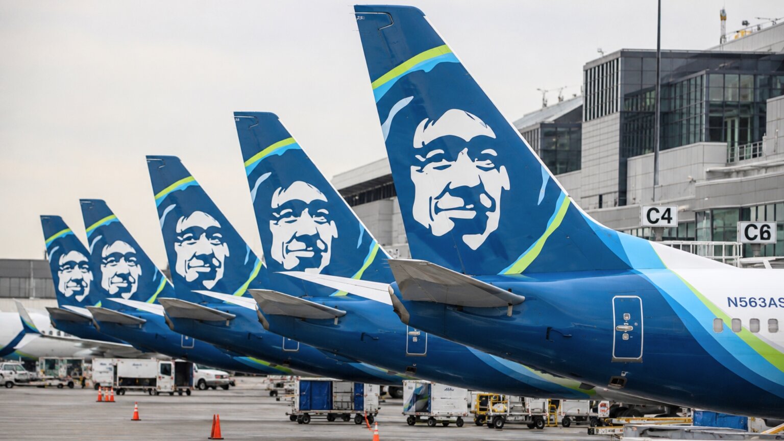 Several Alaska Airlines aircraft parked side by side at an airport gate with their blue tail fins lined up in view.