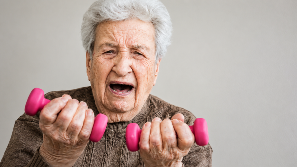 An elderly woman holding small pink dumbbells and grimacing during a workout.