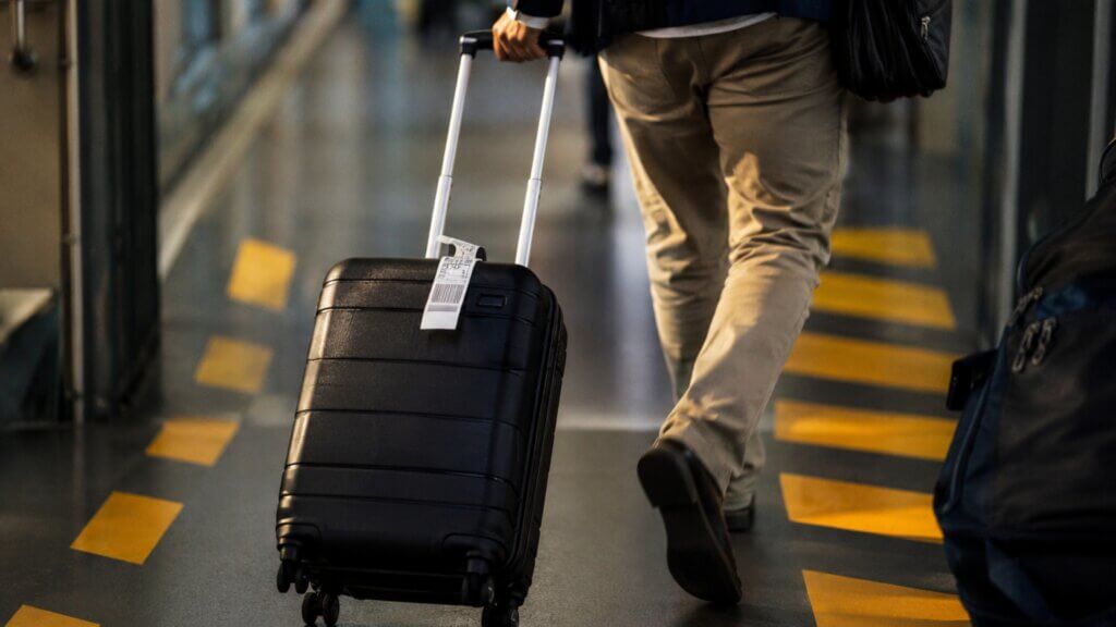 Person walking through an airport terminal while pulling a black rolling suitcase with a baggage tag attached.