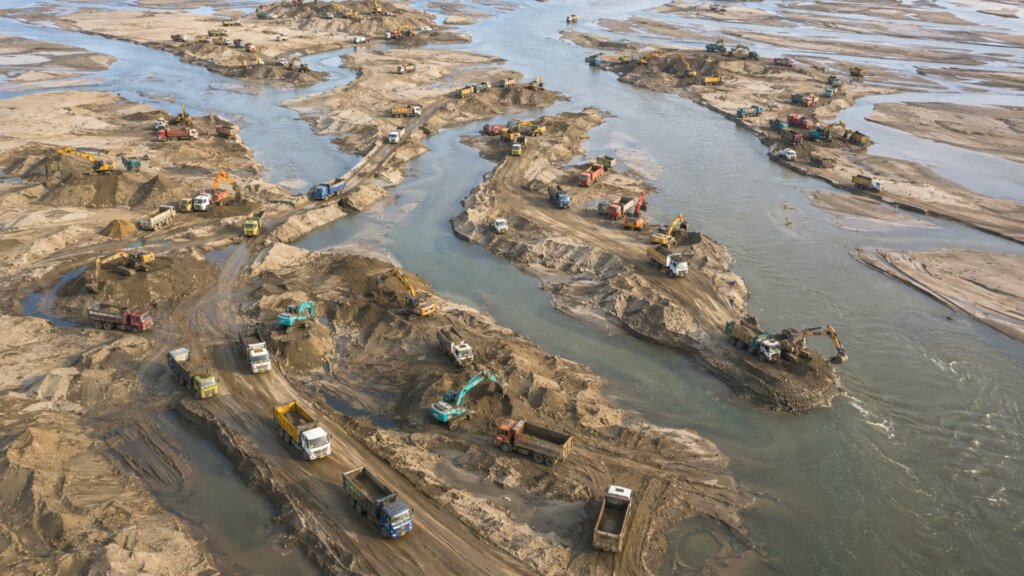 Aerial view of a wide river with multiple sandbars where dozens of excavators and dump trucks are actively excavating and transporting sediment.