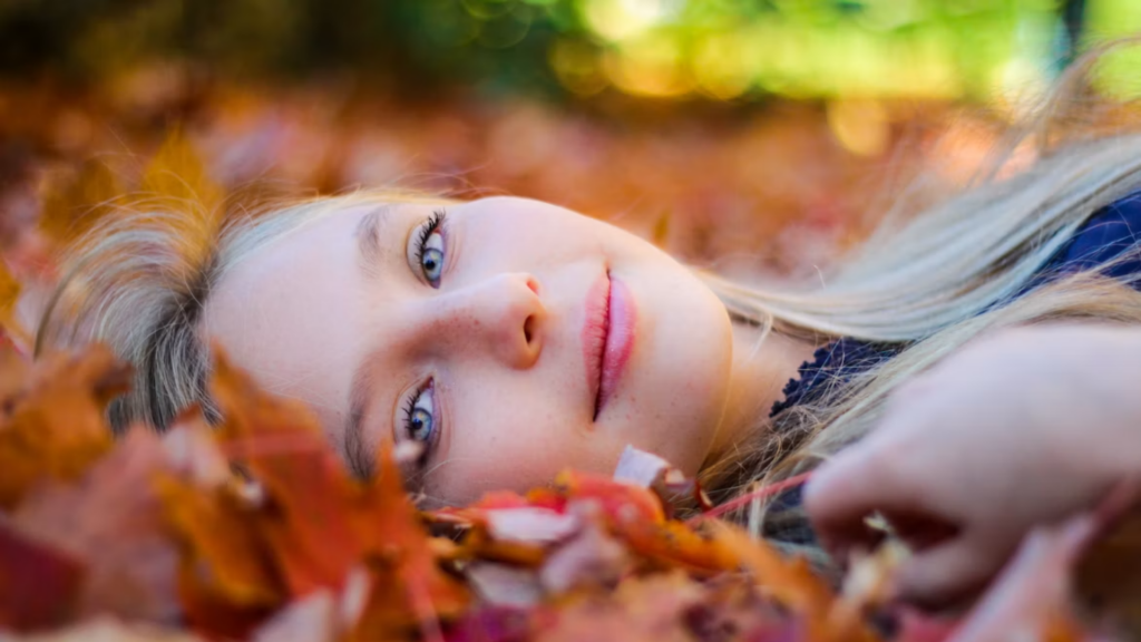Close-up beauty of a woman lying down in autumn leaves with berry-toned lips set against warm autumn light—capturing the season's effortless, lived-in glam look.
