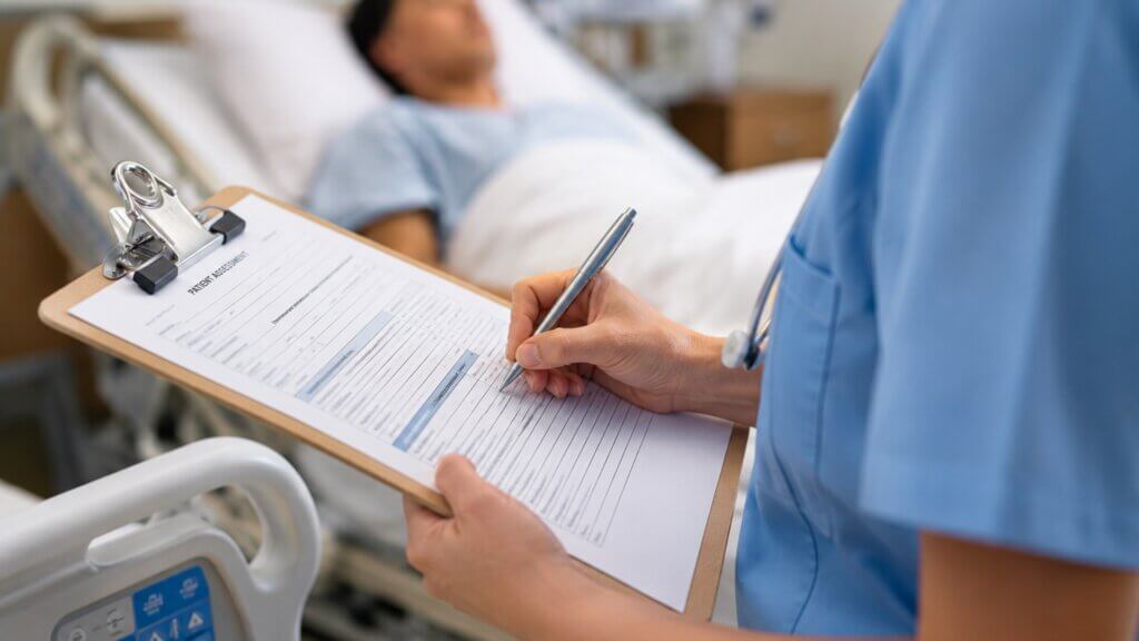 A healthcare worker in blue scrubs writes on a patient assessment clipboard next to a hospital bed where a patient is lying down.