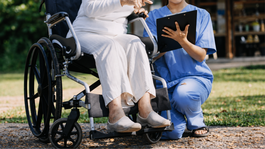 A nurse in blue scrubs holding a clipboard while assisting a patient sitting in a wheelchair.