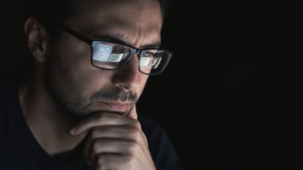 Close up of a man wearing glasses and resting his chin on his hand while looking intently at a screen.