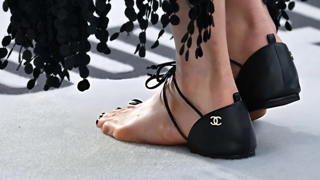 Close-up of black Chanel lace-up heel-cap sandals worn on bare feet with black nail polish.