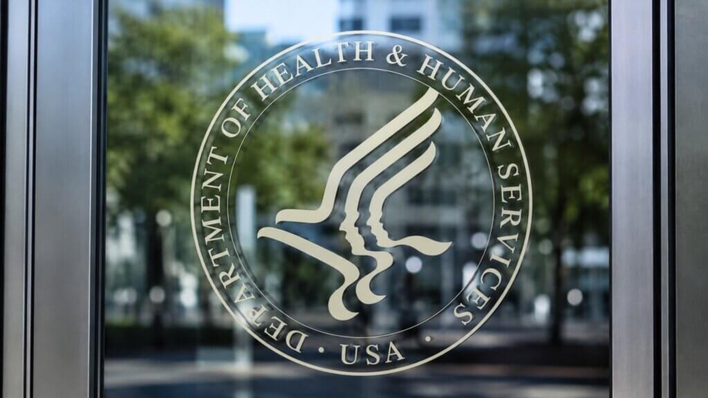 Glass entrance door displaying the seal and text "Department of Health & Human Services USA" with office buildings and trees reflected in the background.