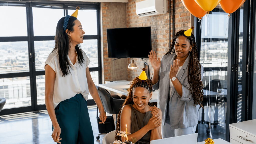 Three coworkers wearing party hats celebrating a birthday with a cupcake in an office.