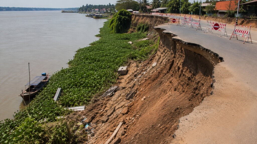 A section of riverside road has collapsed into a steep eroded riverbank, leaving the pavement hanging over the edge.