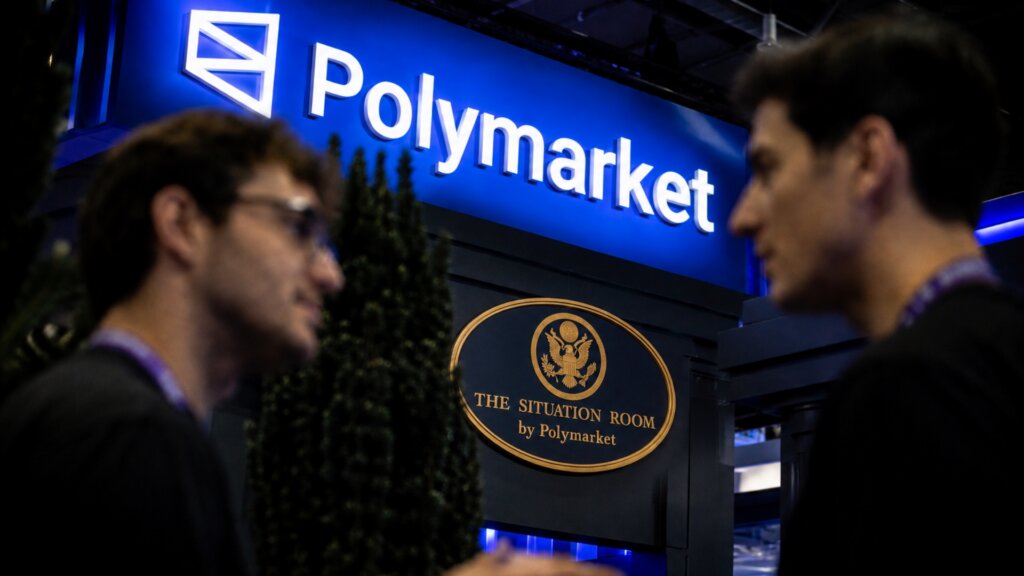 Two people stand in front of a blue Polymarket display.