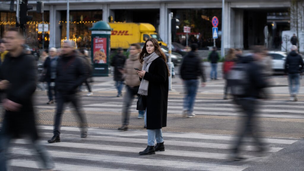 Woman holding a smartphone while standing still at a busy city crosswalk as blurred pedestrians move around her.