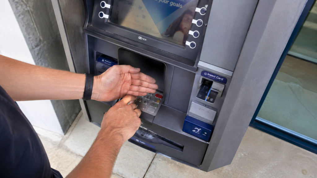 A person covering the keypad with one hand while entering a PIN at an outdoor ATM machine.