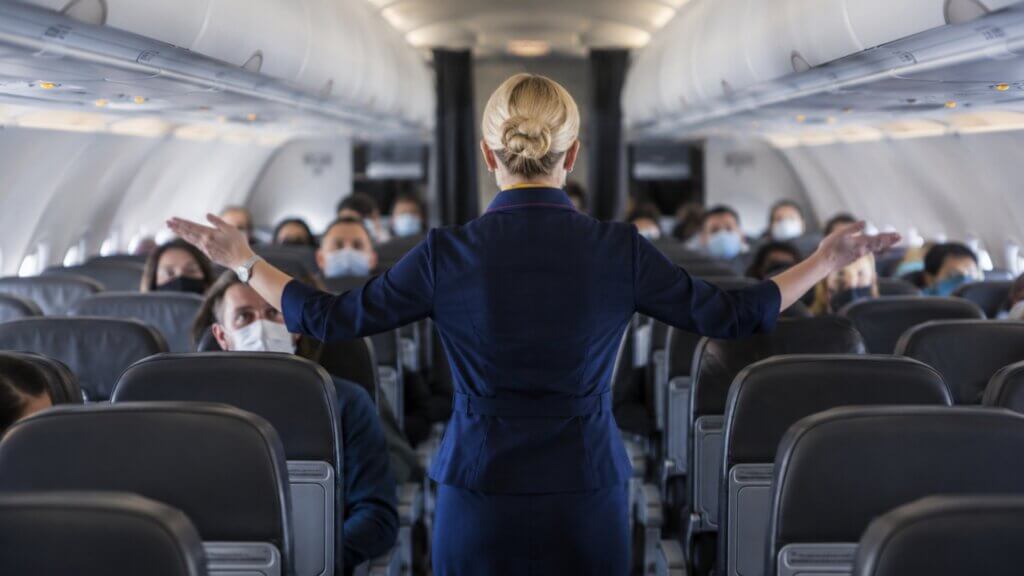 A flight attendant stands in the aisle of an airline cabin with both arms extended while addressing seated passengers.