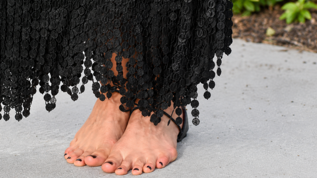 Direct shot of bare feet wearing black lace-up ankle straps under a fringed black dress hem.