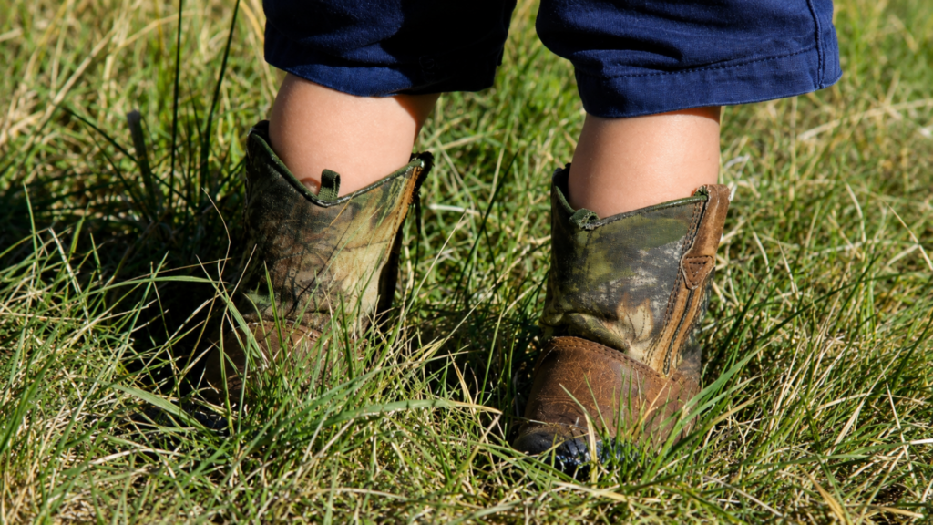 Toddler's feet wearing camouflage and brown leather boots standing in tall grass.