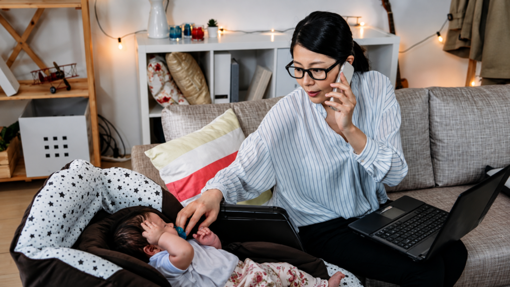 A woman talking on the phone and working on a laptop while tending to her baby in a carrier.
