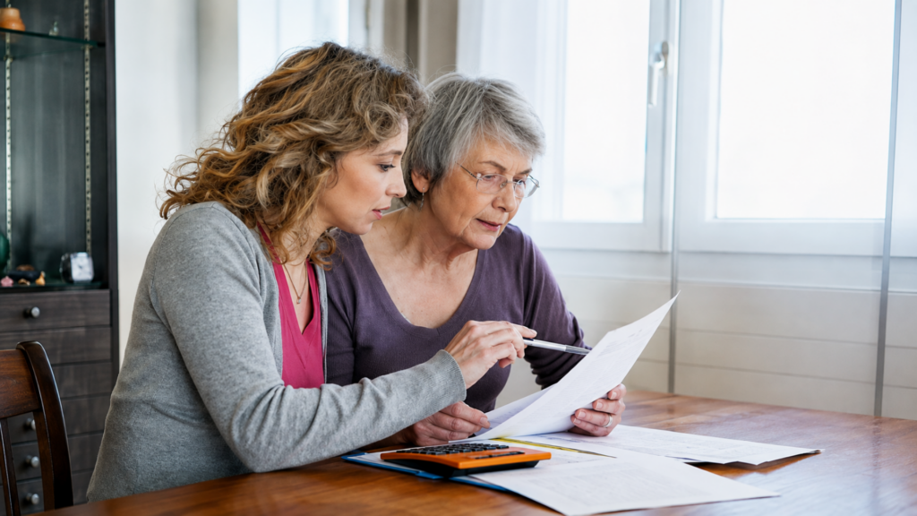 Younger woman helping a senior woman review paperwork with a calculator nearby.