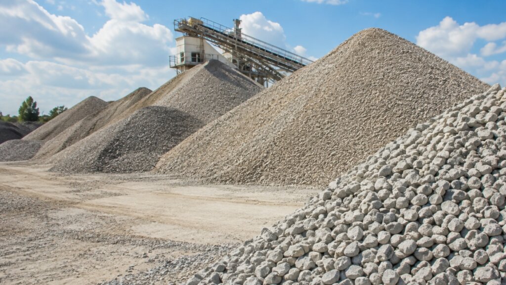 Large piles of sorted gravel and crushed stone sit beside a conveyor system at an aggregate processing facility.
