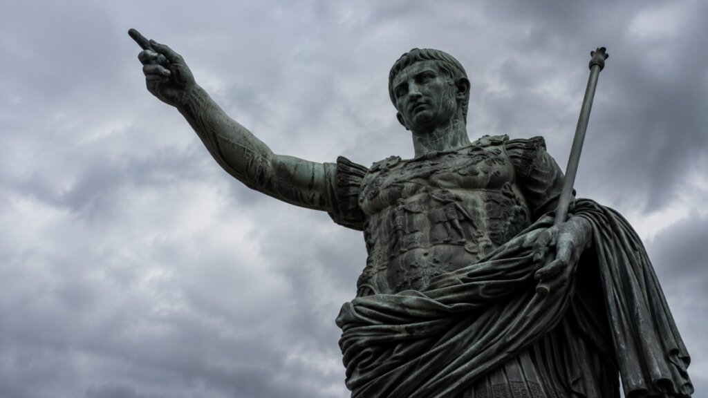 A weathered bronze statue of Julius Caesar stands beneath a cloudy gray sky.