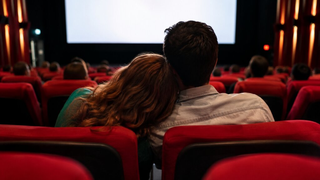 Two people sit close together in red movie theater seats while facing a large screen. One person rests their head on the other persons shoulder as an audience fills the surrounding rows.