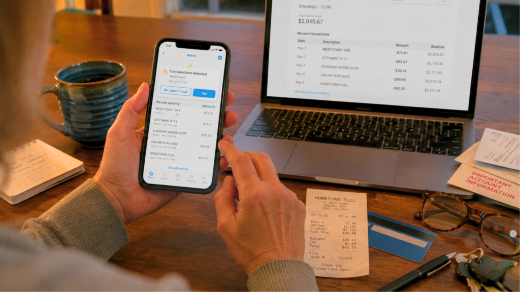 A person reviewing a fraudulent transaction alert on a smartphone next to a laptop displaying online bank activity.