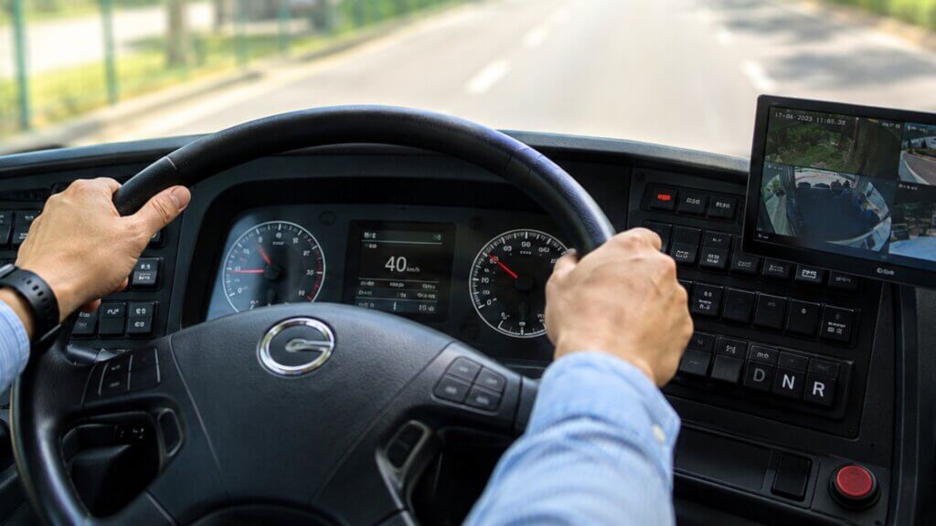 Close up view from the driver's seat of an airport shuttle bus with hands on the steering wheel, dashboard controls, and the road ahead visible through the windshield.