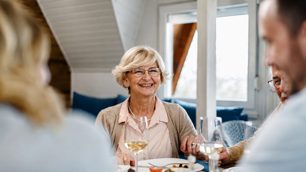 An elderly woman smiling and holding hands with family members at a dinner table.