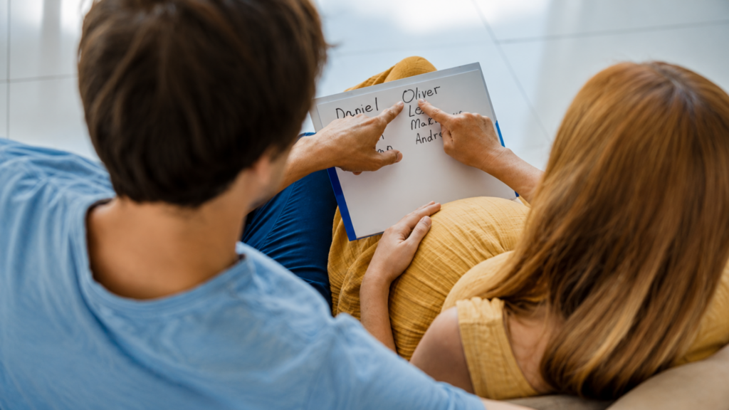 Pregnant couple sitting together looking at a handwritten list of baby names on a whiteboard.