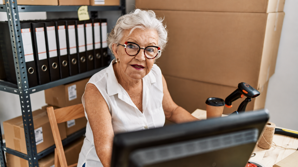 Senior woman wearing glasses working at a computer in a warehouse with shipping boxes.