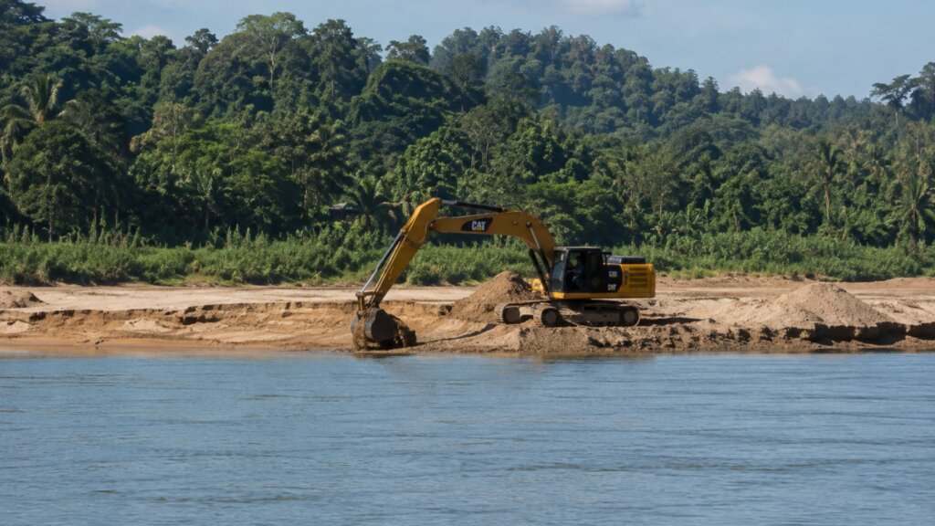 An excavator operates on a sandy riverbank with its bucket extended into the shoreline beside a wide river.