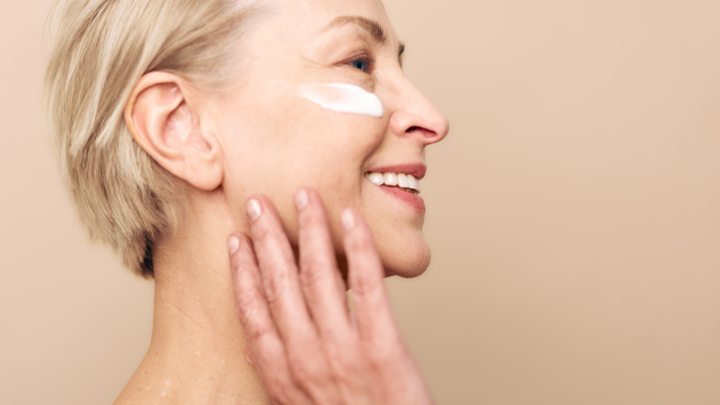 Mature woman smiling while applying skincare cream to her face.