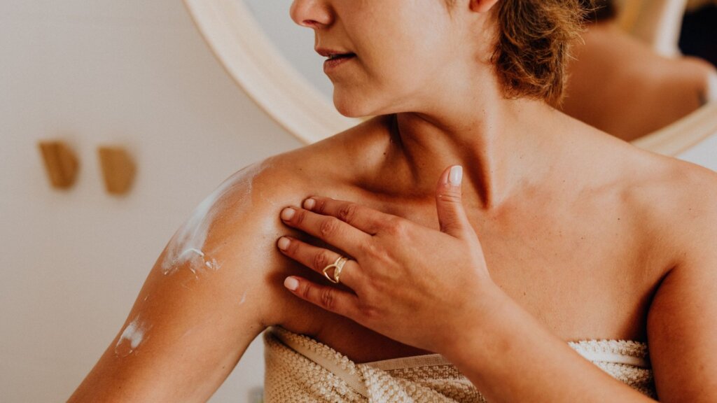 A woman with a towel wrapped around her body applying moisturizer to her shoulder, standing in front of a round mirror.