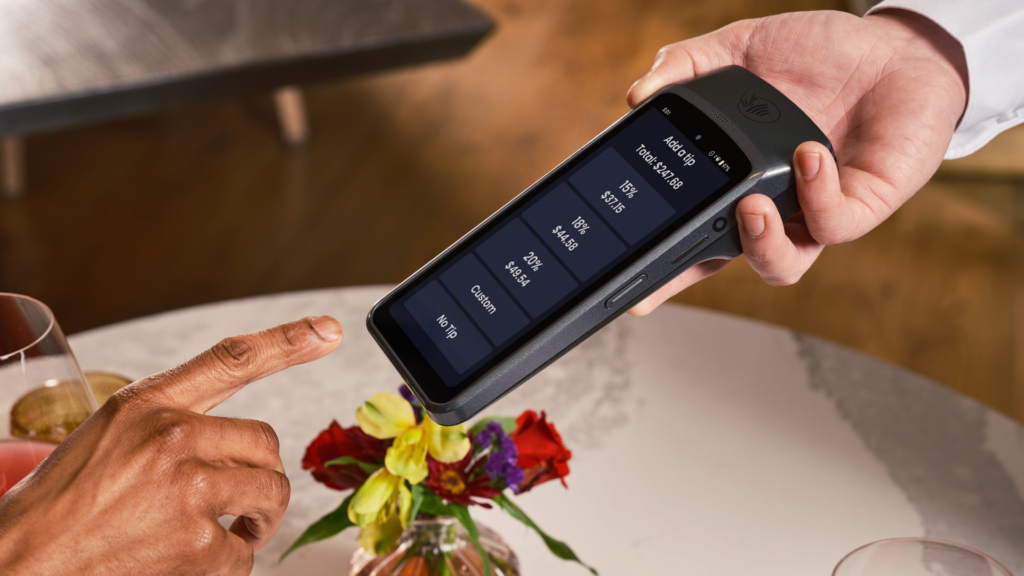 A customer reaches out to tap a tip option on a handheld payment terminal presented by a server at a restaurant table.