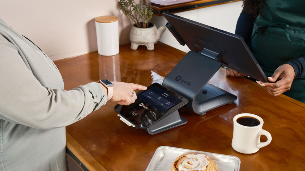 A customer selects an option on a customer-facing payment screen at a bakery counter displaying a cinnamon roll and a mug of coffee.