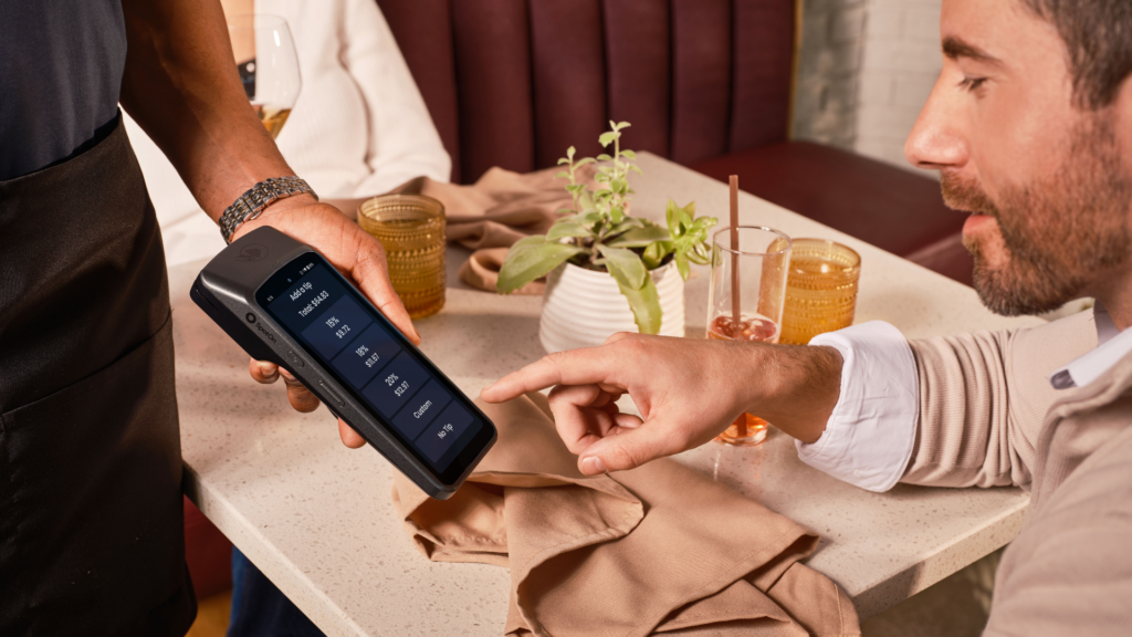 A diner points at the tip screen on a handheld point-of-sale terminal held by a restaurant server.