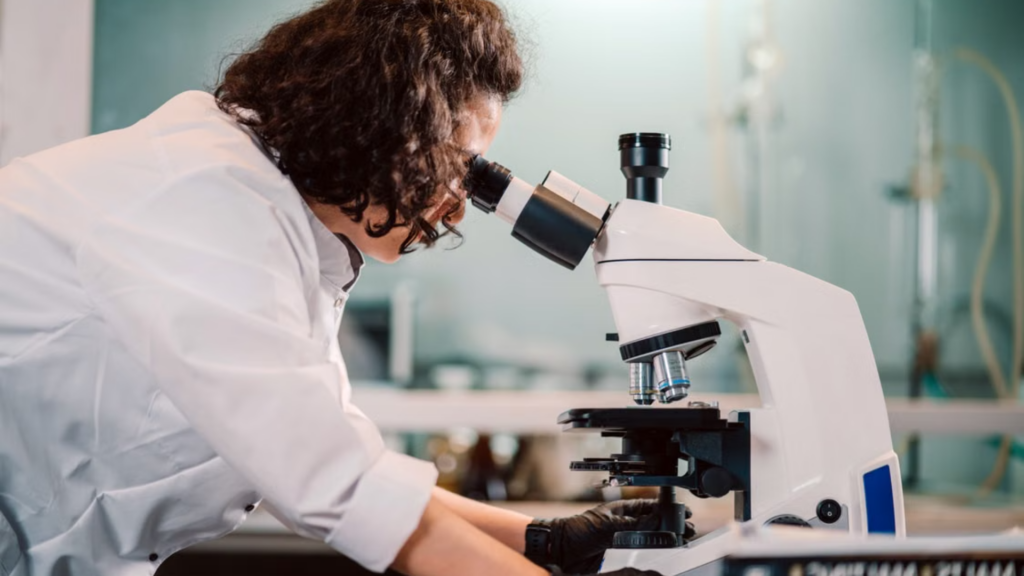 A female scientist in a white lab coat and black protective gloves leans over to examine a sample through a compound microscope in a laboratory setting.