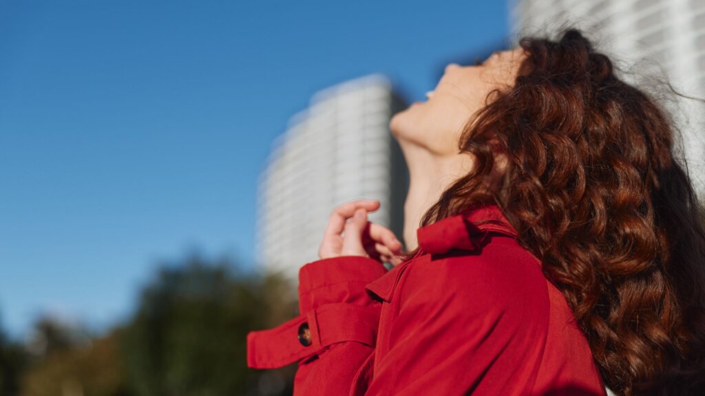 Woman with curly red hair in a red jacket, looking up at the sky with buildings in the background.