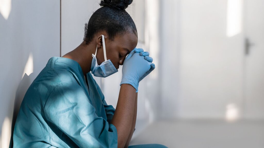 Healthcare worker in scrubs and a face mask, sitting with head bowed and hands clasped against their forehead.
