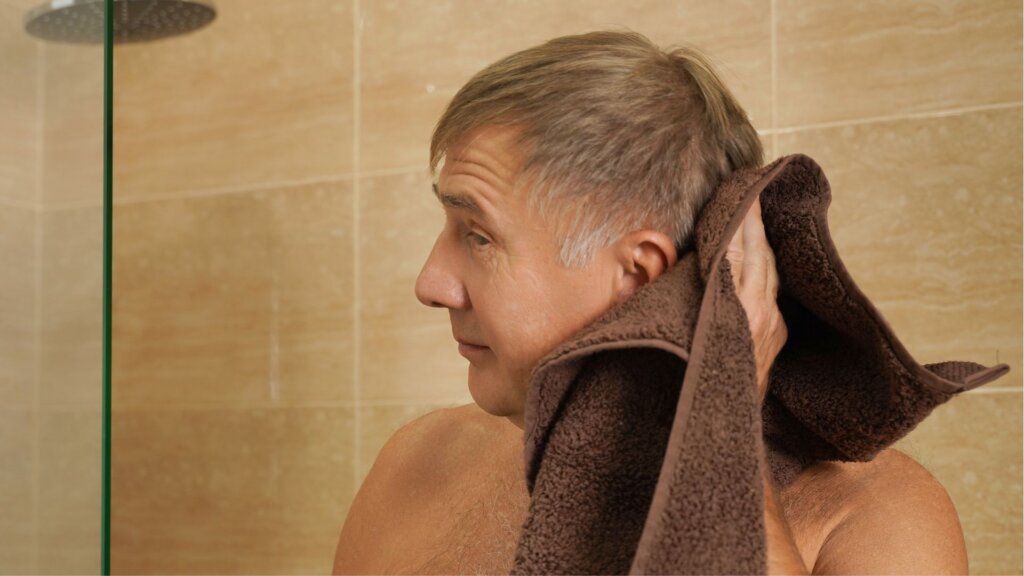 A man with gray hair drying his head with a brown towel while standing near a shower in a tiled bathroom.