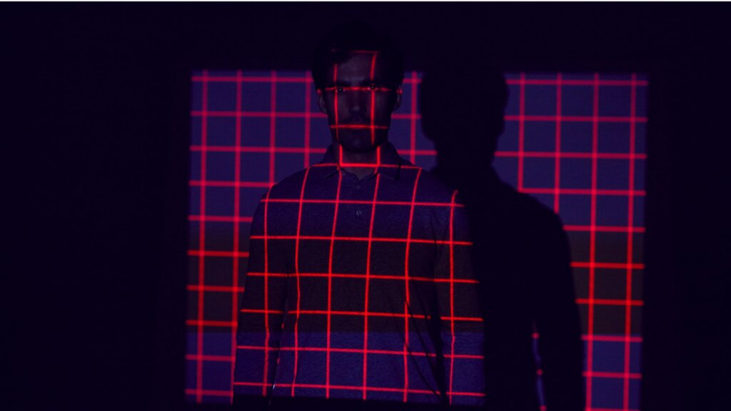 A man stands in a dark room with a red grid pattern of light projected across his face, shirt, and the wall behind him