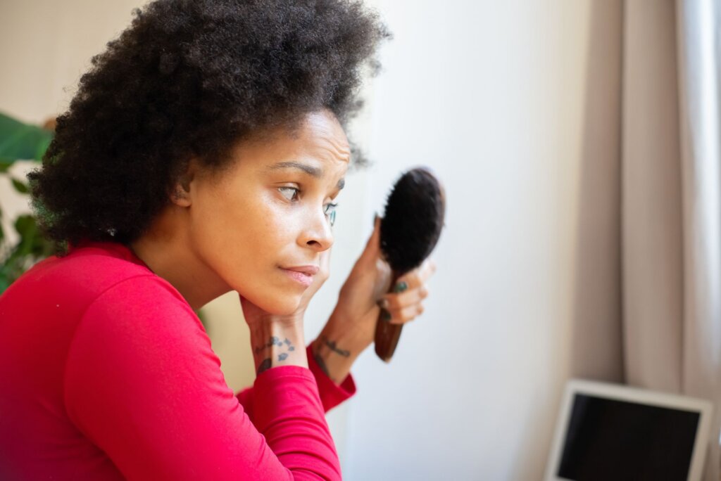 Woman holds a hairbrush while examining her hair near a window.