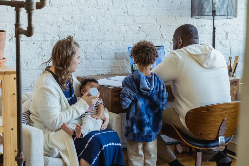 Woman bottle-feeds a baby while a child stands beside a man working at a computer in their home.