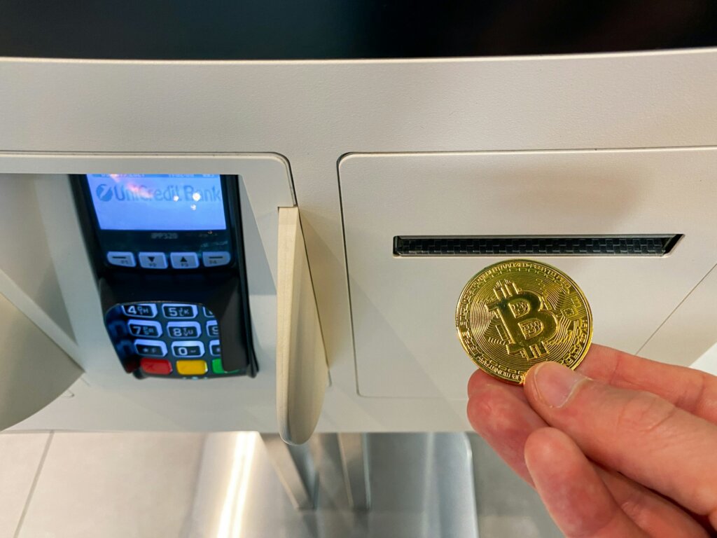 Hand holding a physical Bitcoin token near a cryptocurrency ATM's deposit slot.