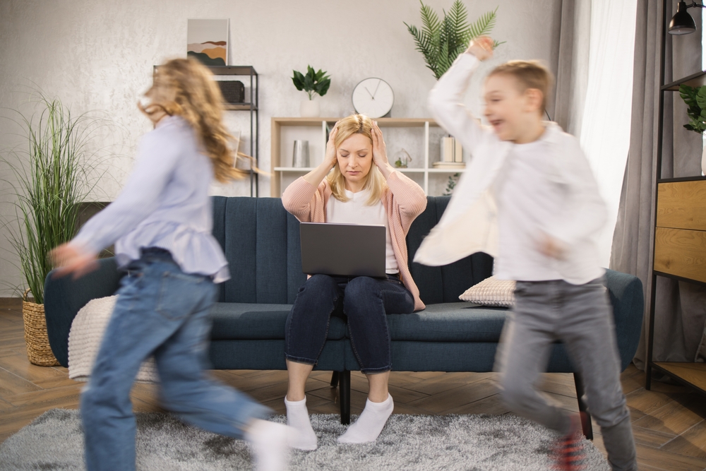 Woman works on a laptop as two children run around her in a living room.